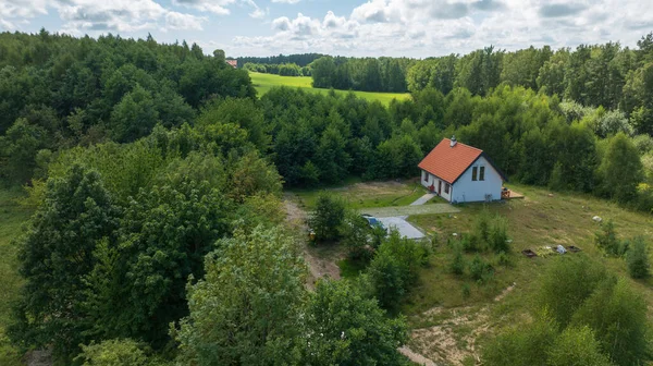 Hava görüntüsü, orman ve çayırların yanındaki kırmızı çatılı küçük beyaz bir ev gösteriyor. Taş bir yol, park edilmiş bir araba, bahçe arsaları ve fidanlıklar yaz bulutlarının altında mevcuttur..