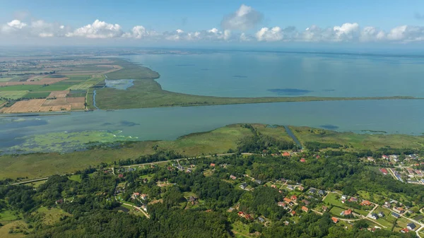 Uzun dar bir tükürük, yamalı tarlalar, sulak alanlarda düz bir kanal ve kıvrımlı bir yol boyunca uzanan sık ağaçlarda kırmızı çatı evleri olan bir kıyı lagününün havadan görünüşü..
