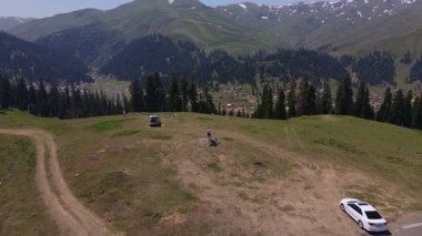 Bir dağ köyü, kozalaklı ormanlar, karla kaplı tepeler, beton bir meydan, iki araba ve yaz ışığı altındaki insanlarla Svaneti, Gürcistan 'da yüksek bir plato manzarası.