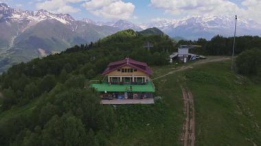 Gürcistan 'ın Svaneti kentindeki kırmızı çatılı dağ kulübesinin havadan görünüşü, teleferik kuleleri, servis binası ve terastaki küçük figürler, kar örtülü Kafkasya zirvelerine kurulmuş..