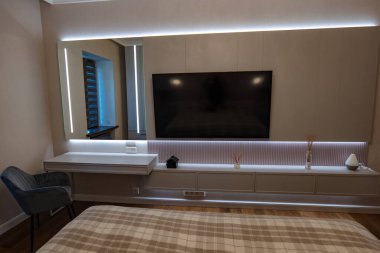 Çağdaş yatak odası bej bir panelde düz ekran bir televizyon, ışıklı ayna ve gri kadife sandalyeli yüzen bir masa ve alevli bir dolap gösteriyor..