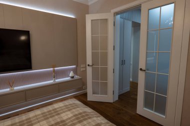 Düz bir gösteride bej yatak odası, LED aksanlar, dolapta inşa edilmiş, duvar TV, ekose yorgan, koridordaki gardıroplara açılan soğuk Fransız kapıları, sıcak ahşap zemin, minimalist dekor..