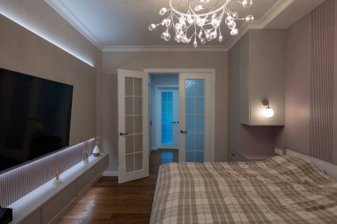 Bir dairedeki çağdaş yatak odası cam küreli bir avize, bir duvar lambası, ekose yatak, LED ışıklandırmalı medya konsolu ve akşamları çift cam kapılar gösteriyor..