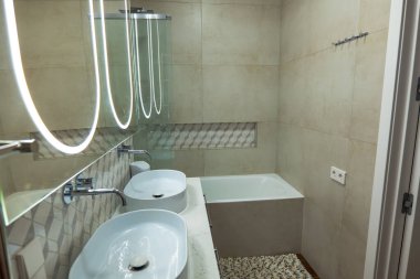 Kompakt banyo oval damar lavaboları, krom musluklar ve halka, bej fayanslar ve küvetin üzerinde desenli bir desenli neon ışıklı büyük bir arka ayna ile çift kibir gösteriyor..