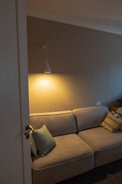 Kısmen açık bir kapı aralığı, desenli yastıklar, beyaz bir duvar lambası, bej desenli bir duvar, kartonpiyer, prizler, kutular ve akşam ruh hali ile boz bir bölüm gösterir..