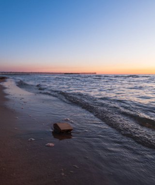 Nazik dalgalar, Litvanya 'nın Palanga şehrinde alacakaranlıkta kumlu bir sahile vurur. Palanga İskelesi şeftali ve mavi gökyüzünün altında ufuk boyunca uzanırken, küçük bir akıntıya kapılmış ağaç parçası ıslak kumun yanında duruyor..