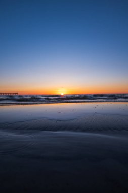 Litvanya 'daki Baltık Denizi üzerinde bir iskele sola doğru uzanırken güneş batar. Cam gibi bir gelgit havuzu turuncu ufku ve mavi gökyüzünü yansıtır. Düz kum ve alacakaranlıkta hafif dalgalar..