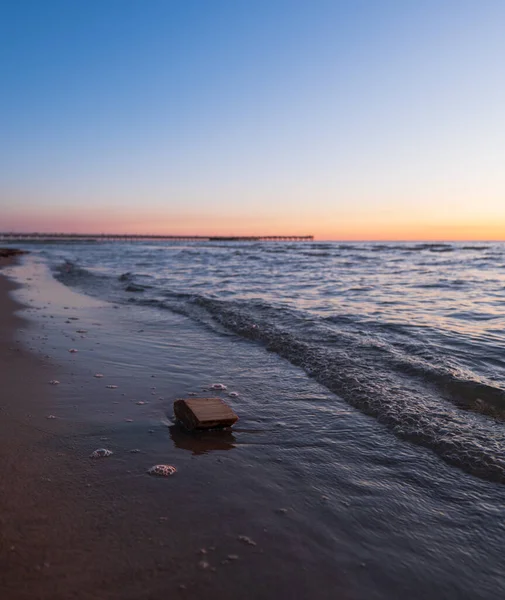 Nazik dalgalar, Litvanya 'nın Palanga şehrinde alacakaranlıkta kumlu bir sahile vurur. Palanga İskelesi şeftali ve mavi gökyüzünün altında ufuk boyunca uzanırken, küçük bir akıntıya kapılmış ağaç parçası ıslak kumun yanında duruyor..