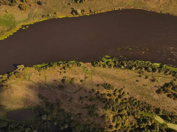 Hava manzarası Litvanya 'nın kırsal kesimlerinde kıvrılan geniş, karanlık bir nehir gösteriyor. Sazlıklar, dağınık ağaçlar, güneşli çayırlar, ağaçlık yollar ve sol altta küçük bir gölet..