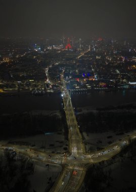 Aerial night scene in Warsaw shows Swietokrzyski Bridge over Vistula River, light trails on snowy roads, and the Palace of Culture and Science glowing red amid hazy towers.