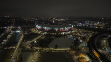 Hava gece görüşü, Varşova PGE Narodowy 'nin Vistula Nehri' nin kıyısında kar, kavis çizgileri, demiryolu hatları, şehir ışıkları ve kırmızı bir çatı kulesi ile kırmızı ve beyaz parladığını gösteriyor..