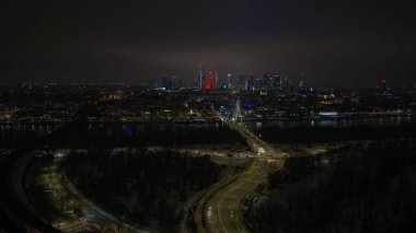 Aerial night scene in Warsaw shows Swietokrzyski Bridge over the Vistula, the Palace of Culture and Science glowing red, curving highways, and low clouds with neon accents.