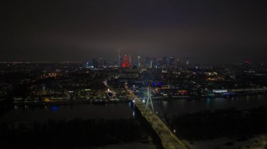 Aerial night scene of central Warsaw shows Swietokrzyski Bridge over the Vistula River, the Palace of Culture and Science in red, Warsaw Spire, and Varso Tower under clouds.