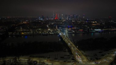 Aerial night view of central Warsaw shows Swietokrzyski Bridge over the Vistula River, the Palace of Culture and Science in vivid red, car lights, neon, and winter snow patches.