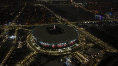 Varşova PGE Narodowy 'nin kırmızı ve beyaz parlayan kış gecesi manzarası, Vistula Nehri ve köprüler pırıl pırıl, karlı yürüyüş yolları, ufuk boyunca yayılmış şehir ışıkları.