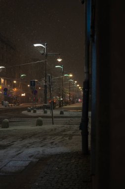 Geceleri eski sokak lambalarının altında tozlu bir şehir sokağı kıvrımları oluşuyor. Düşen taneler sıcak ışığı yakalar, tramvay telleri ve kalın ipler yürüyüşü kaplar ve buzlu asfalt yansıtır..