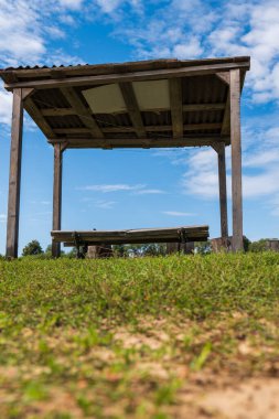 Parlak mavi, parçalı bulutlu gökyüzünün altında, gün ışığında sığ bir odak noktasıyla çekilmiş, eğimli çatılı bir bankın üzerinde duran kırsal bir ahşap barınak..