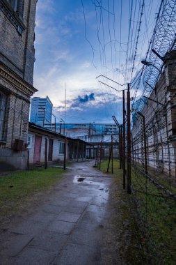 Dikenli teller ve tuğla duvarlar arasında dar nemli bir yürüyüş yolu var, bahçeye çıkıyor. Uzun dikenli tel duvar, cam gibi yüksekte dikiliyor..