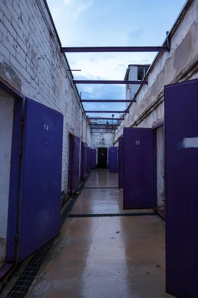 Vilnius 'taki Lukiskes Hapishanesi' nin dış hücre koridorunda 14 ve 22 numaralı aralık mor kapılar görünüyor. Kırık duvarlar, ıslak beton ve bulutlu akşam ışığı..