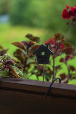 Sığ bir derinlik görüntüsü, kırmızı yapraklı ahşap bir balkon ekicisi ve Litvanya 'da bir bahçe sahnesi olan PODLEJ NAS yazan küçük bir ev şeklindeki kazığı gösteriyor..