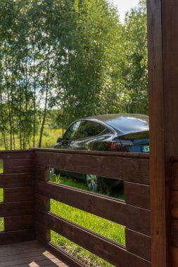 Litvanya 'nın kırsal kesimindeki ince huş ağaçlarının yanındaki çimlerin üzerinde parlak siyah bir elektrikli araba çerçeveleniyor. Sakin bir yaz sahnesinde, öğleden sonra ışığı çatıdan yansıyor..