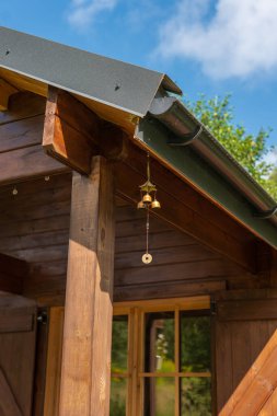 Litvanya kırsalındaki ahşap kulübe verandasında koyu yeşil metal çatı, yağmur oluğu ve pirinç rüzgar çanı görülüyor. Sıcak kereste, kepenkli pencere, yaz ışığı, mavi gökyüzü, yumuşak bulutlar..