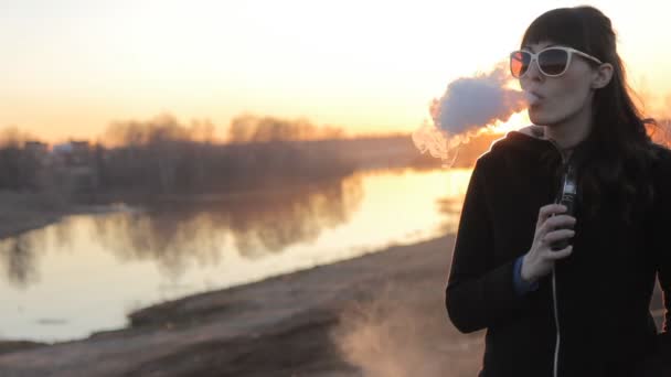 Chica en un fondo del río fuma un cigarrillo electrónico — Vídeo de stock