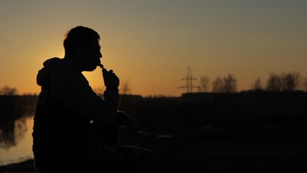 Um jovem ao pôr do sol fumando cigarro eletrônico — Vídeo de Stock