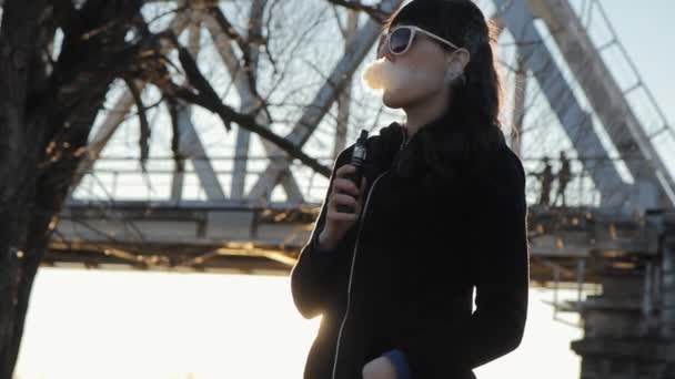Meisje op de achtergrond van de brug rookt een elektronische sigaret, dolly — Stockvideo