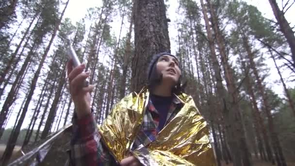 Lost female tourist wrapped in a golden survival blanket in the woods catches a connection on her smartphone — Stock Video