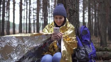 Altın orman battaniyesine sarılı pozitif kadın turist akıllı telefon kullanıyor.
