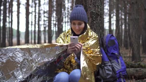 Позитивна жінка-турист, загорнута в золоту лісову ковдру, використовує смартфон — стокове відео