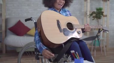 Tekerlekli sandalyede afro saç stili olan engelli Afrikalı Amerikalı kadın akustik gitar çalıyor.