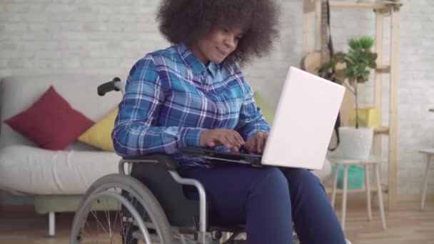 Mujer afroamericana discapacitada con un peinado afro en una silla de ruedas utiliza una computadora portátil mirando a la cámara — Vídeos de Stock