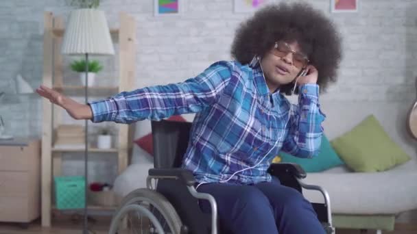 Mujer africana discapacitada alegre con un peinado afro en una silla de ruedas en auriculares escuchando música y cantando — Vídeo de stock