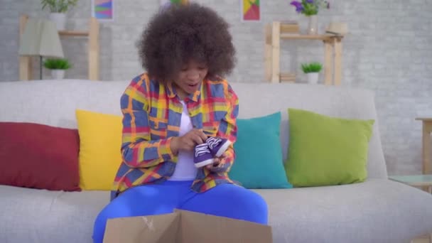 Alegre y feliz mujer afroamericana con un peinado afro desempaca el paquete, sentado en el sofá — Vídeo de stock