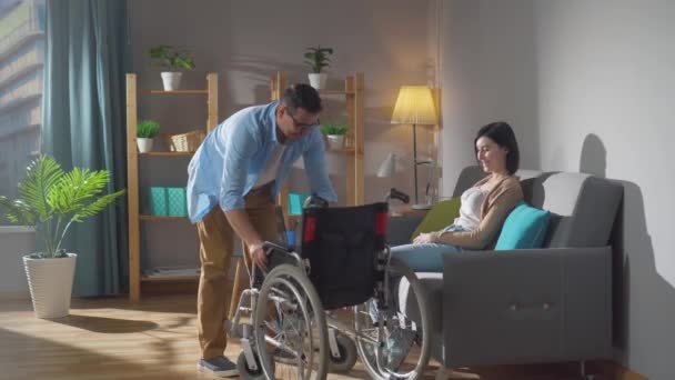 Pareja de cónyuges una joven discapacitada y su marido la ayudan a entrar en una silla de ruedas — Vídeo de stock