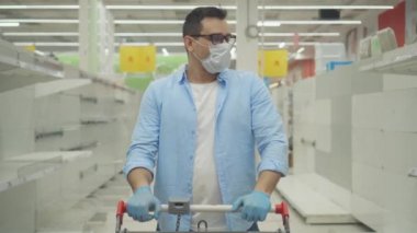 Boş rafların arkasında maske ve eldiven takan bir adam.