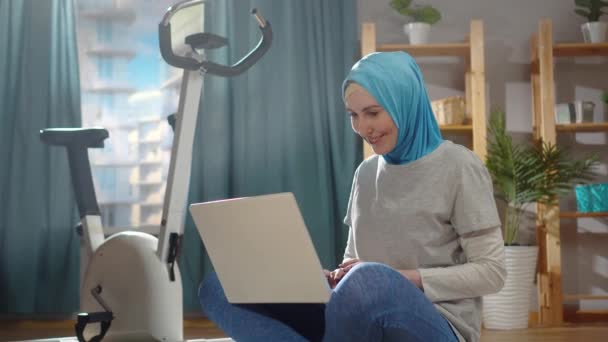 Deportiva mujer musulmana en un pañuelo nacional en la sala de estar con un ordenador portátil tirado en el suelo — Vídeos de Stock
