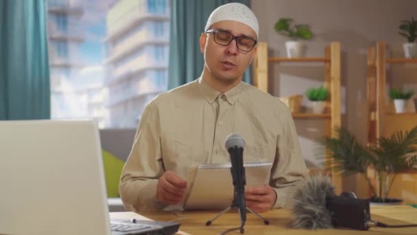 Porträt muslimischer Mann mit Totenkopf nimmt einen Podcast auf, während er im Wohnzimmer vor einem Mikrofon sitzt — Stockvideo