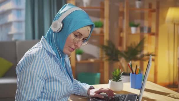 Retrato jovem muçulmano freelancer em um lenço de cabeça nacional trabalha em um laptop sentado na sala de estar — Vídeo de Stock