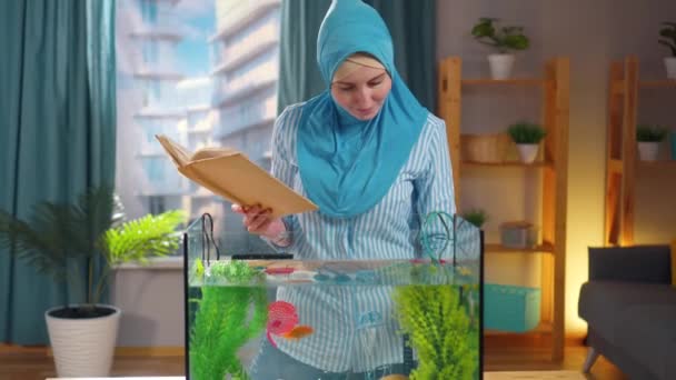 Mulher muçulmana em lenço de cabeça nacional estuda um livro sobre aquários e alimenta peixes dourados em um aquário — Vídeo de Stock