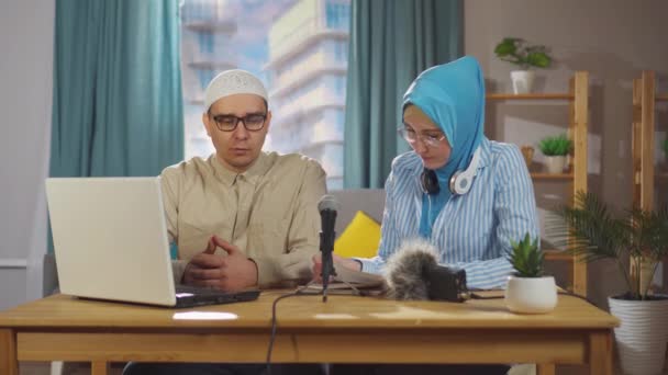 Ung muslimsk kvinna och en muslimsk man spelar in en intervju podcast när de sitter framför en mikrofon i vardagsrummet — Stockvideo