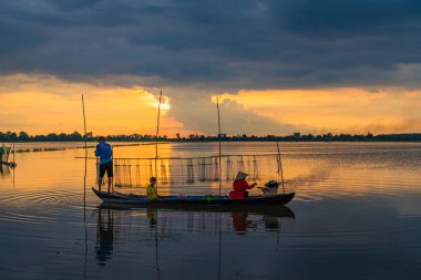 Long An, Vietnam - 22 Ağustos 2024: güzel, huzurlu bir gün batımı. Yerel bir aile, bir baba, bir çocuk ve bir anne küçük ahşap bir teknede ağlarıyla ilgilenirken ya da güneş ufkun altına batarken balık tutarken görülürler.