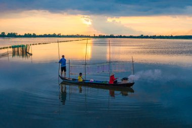 Long An, Vietnam - 22 Ağustos 2024: güzel, huzurlu bir gün batımı. Yerel bir aile, bir baba, bir çocuk ve bir anne küçük ahşap bir teknede ağlarıyla ilgilenirken ya da güneş ufkun altına batarken balık tutarken görülürler.
