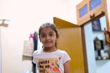 Siyah saçlı bir kız açık kapılı bir odada duruyor. Gülümsüyor ve tişört giyiyor..