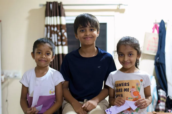 Üç çocuk, ellerinde renkli el sanatları ile akşam boyunca bir ev ortamında gülümsüyor..