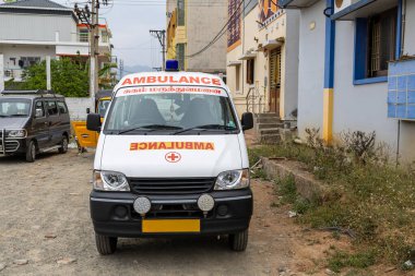 Tozlu bir caddeye park etmiş bir ambulans gündüz saatlerinde binalar ve araçlarla çevrili..