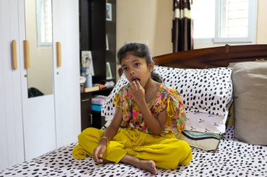 Bir çocuk evde otururken yatakta oturur ve atıştırmalık şeyler yer..