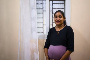 Hamile kadın pencereden dışarı bakar, rahat bir evde beklenti ve neşe saçar..